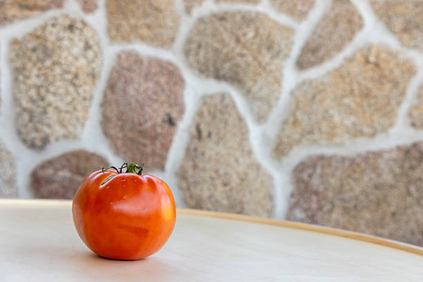 A tomato sits on a table in front of a stone wall. The tomato is ripe and ready to be eaten