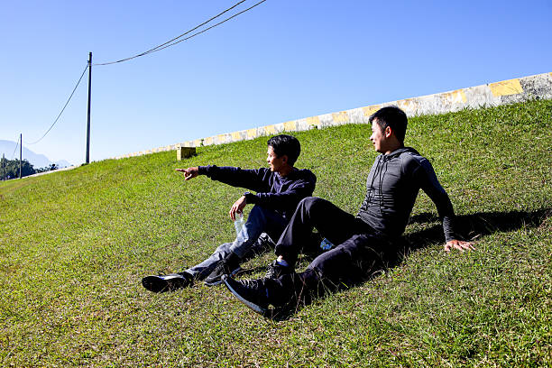 Two Asian Men Relax On A Grassy Hill, Enjoying A Sunny Day Outdoors
