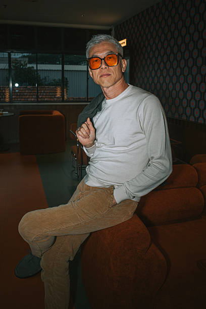Portrait of a silver-haired Asian man in casual fashion, wearing tinted glasses and leaning back confidently in a retro-themed lounge. He sits on a brown velvet armchair with one hand in his pocket and a relaxed expression, conveying timeless style and ease. The interior features mid-century modern decor and warm tones. Shot in Malaysia.