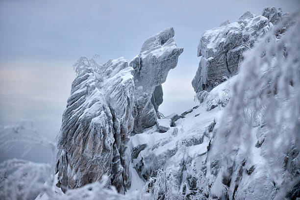Unique rocky formation draped in thick snow creates a striking contrast against cloudy winter sky. Frosty trees surround area, enhancing the serene and chilly atmosphere. Taganay Nature Park, Russia