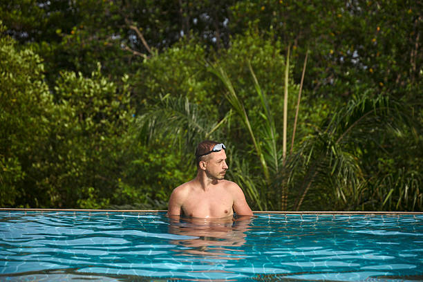 shirtless man is sitting in an infinity pool, enjoying the calm water while surrounded by vibrant green foliage. sunlight reflects off the pool, creating a serene atmosphere.