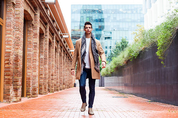 Stylish young Caucasian man walking confidently on a brick path between old brick walls and a modern glass building, wearing a camel coat and scarf.