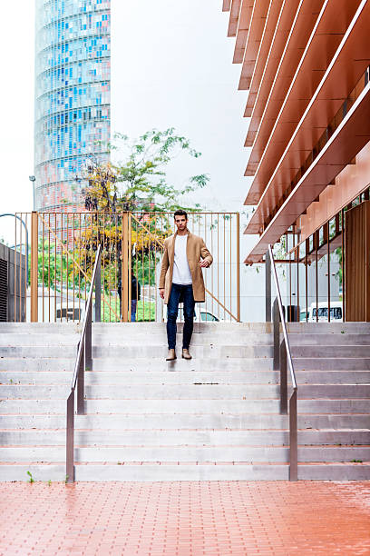 Young Caucasian man in a camel coat, jeans and boots walking down wide steps in a modern urban area, with contemporary buildings in the background.