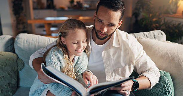 istock Father, girl and reading book on couch, learning and support with development, love and bonding in lounge. People, dad and child with literature, education and help with smile on sofa at family house 2228023786
