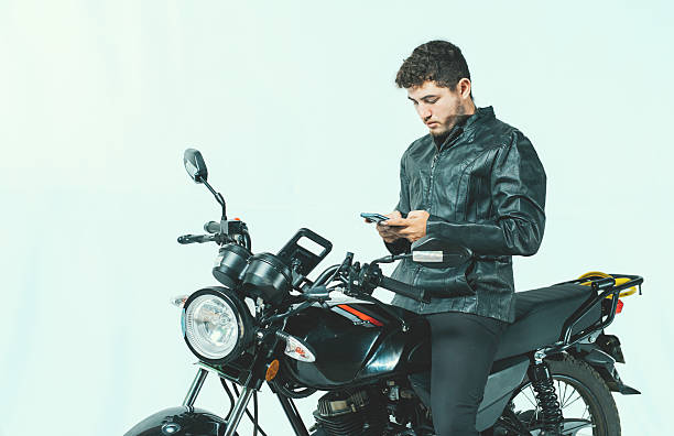 istock Young motorcyclist using his cell phone while sitting on his motorcycle, isolated. Male biker on a motorcycle using his phone isolated on white background 2228026098