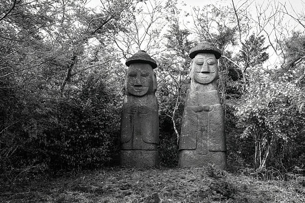 Two traditional Dol Hareubang statues in black and white, located in a forest on Jeju Island, South Korea.