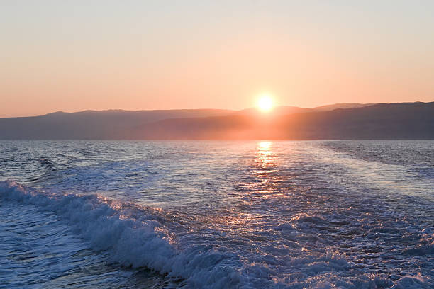 Calm sea at sunset with soft waves and distant hills, glowing in warm tones of orange and purple under a clear sky.