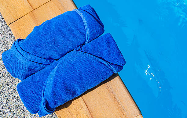 Back View Of A Senior Man With Gray Hair Sitting By A Pool, Wrapped In A Bright Striped Towel. The Scene Captures A Relaxing Moment In The Sun After Swimming, With Greenery And Lounge Chairs In The Background. Concept Of Rest, Wellness, And Healthy Aging.