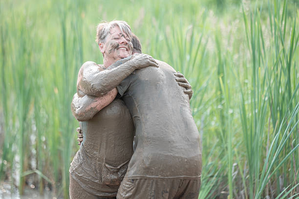 Smiling individuals engage in a joyful embrace, completely covered in mud, surrounded by lush greenery. The image captures themes of fun, connection, and the natural world in an expressive and heartwarming moment.