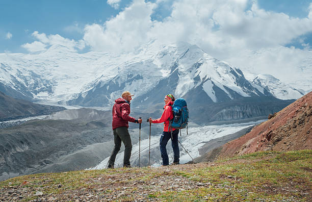 istock Smiling each other backpackers couple with backpacks, trekking poles during Lenin peak ascent with mountain peaks in background. Extreme active people, high-altitude Pamir area mountaineering concept 2228091145