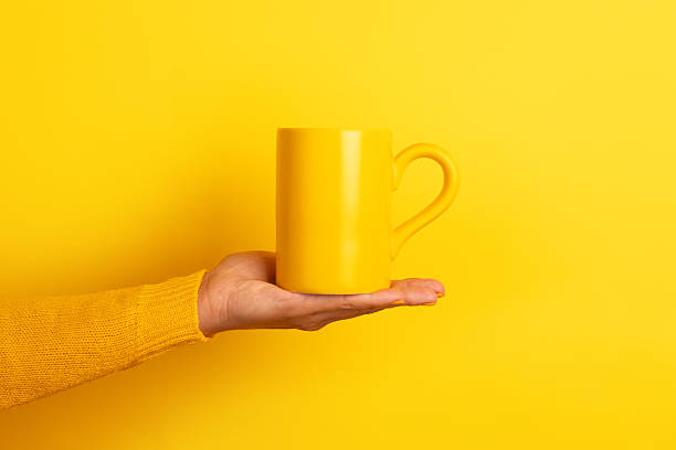 istock Hand in yellow sweater presenting a plain yellow ceramic mug on an open palm against a bright yellow background. 2228094370