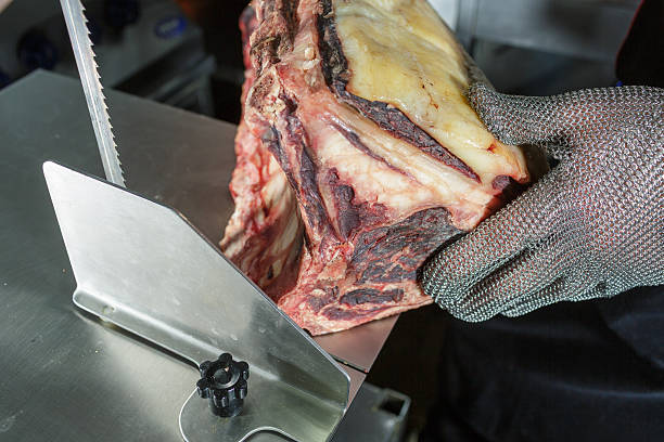 Butcher Slicing Dry Aged Beef with Meat Saw in Kitchen Using Safety Glove on Stainless Table