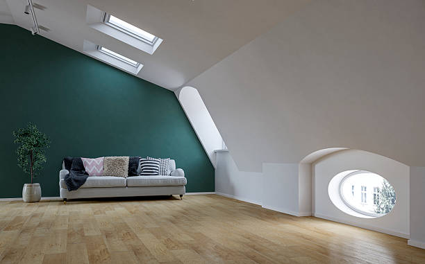 Contemporary attic living room with a striking pine green accent wall and sloped ceilings featuring skylights. A light grey upholstered sofa decorated with a variety of patterned cushions and a soft throw adds warmth to the space. Natural light fills the room through modern architectural windows, including a distinctive oval-shaped window and skylights. The interior is complemented by a light wood floor and a potted indoor plant, creating a cozy yet refined atmosphere. Perfect for use in interior design showcases, real estate, editorial content, or home lifestyle advertising. 3D rendered image.