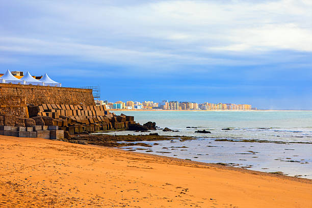 Cadiz shoreline of Atlantic Ocean unfolds with a sandy beach, rugged seawall, and white tents perched above the concrete blocks. Cityscape stretches behind, framed by a moody, cloud sky
