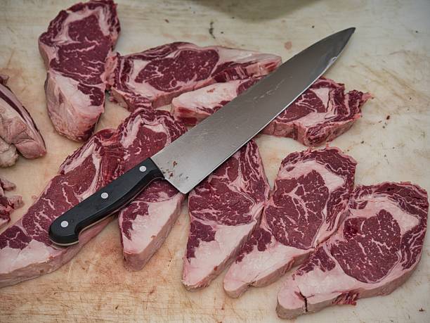 A close-up view of raw beef steak served on a ceramic plate in a restaurant. A chef's hand with a knife slices the fresh meat before cooking. Realistic food photography showing traditional meal preparation with natural light and authentic setting.