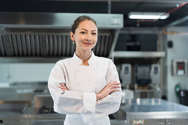 istock Restaurant kitchen. Female chef standing with crossed arms in kitchen, wearing white apron and smiling looking to camera 2228143031