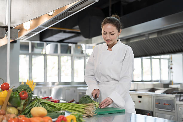 istock Restaurant kitchen. Female chef preparing meal in kitchen, cutting board with vegetables, wearing white apron. Female cooks preparing food in kitchen at restaurant 2228143857