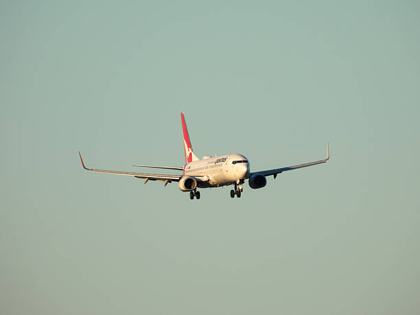 A Qantas Boeing B737-838 plane, registration VH-VZC, approaching to land on the east-west runway of Sydney Kingsford Smith Airport as flight QF452 from Melbourne. This image was taken from a public carpark at the domestic terminal on a cold, windy and sunny afternoon nearing sunset on 11 July 2025.