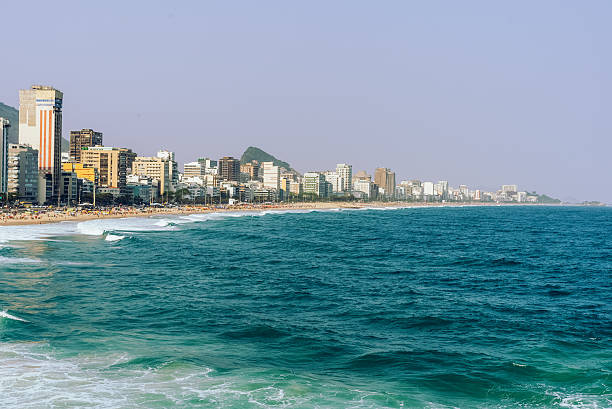 Sunny Rio de Janeiro beach with green lounge chairs in the foreground, crowds along the shore, and dramatic mountains in the distance. Bright sky, surfers, and waves create a lively coastal scene.