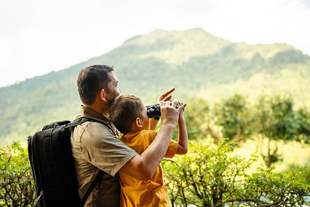 Father showing something to son looking through binoculars at forest during sunny day
