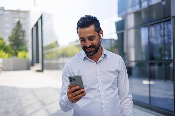 istock Mid adult manager walking confidently while using a smartphone, enjoying music through wireless earbuds in front of modern office buildings 2228155107