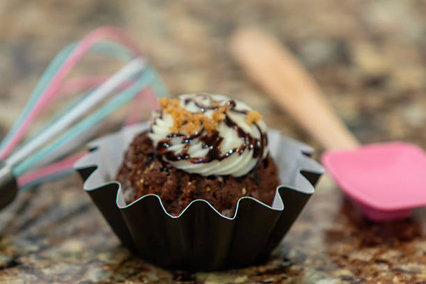 A single-serving mini bundt cake topped with a swirl of cream cheese frosting, presented in a dark fluted wrapper and placed on a polished granite countertop. This close-up food image showcases the cake’s golden texture and elegant decorative icing, suitable for themes related to desserts, celebrations, and home baking.