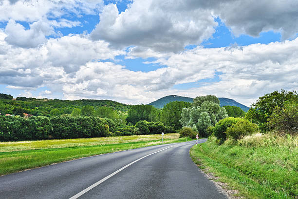 Curved asphalt highway with white lane markings descending through lush green countryside toward forested mountains under dramatic cloudy sky.