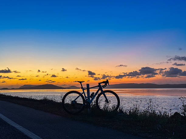 A captivating scene featuring a bicycle's silhouette resting by a serene waterfront during a vivid and picturesque sunset. The sky transitions from warm oranges to deep blues, reflecting beautifully on the water's surface. Perfect for themes related to outdoor recreation, tranquility, and the beauty of nature's quiet moments.