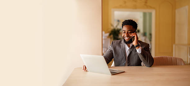 istock Smiling young businessman discussing over mobile phone while using laptop at desk by wall in modern office 2228169406