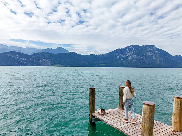 Young woman in casual clothes standing on wooden pier looking at calm turquoise lake. Scenic mountain landscape with cloudy sky in background. Outdoor nature and travel concept.