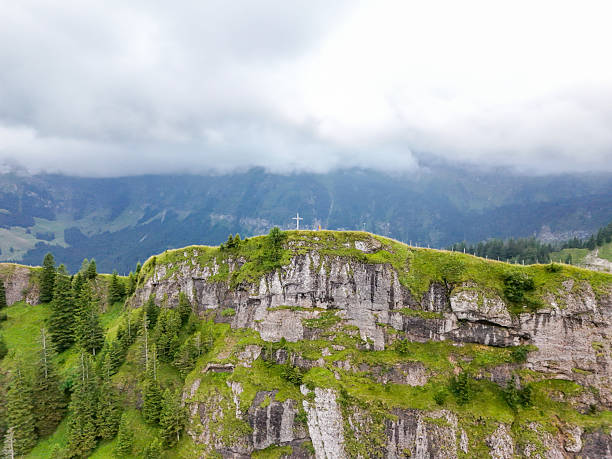 Elevated rocky hill covered with green grass and a white cross on top. Dense pine forest surrounds the hill with misty mountains in the background. Scenic natural landscape with religious symbol.