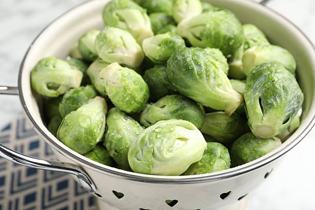 Fresh Brussels sprouts in metal colander on white marble table, closeup