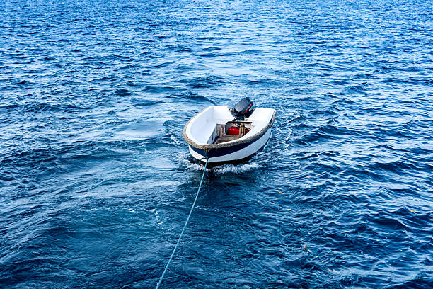 Small motorized dinghy trails gently on the open sea, tethered and drifting through rippling blue waves under clear skies.