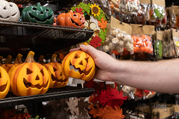 Male hand selecting a classic orange jack-o'-lantern decoration from a crowded retail store shelf, highlighting popular Halloween themed products for festive autumn shopping