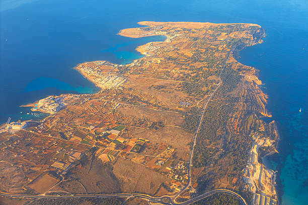 Aerial view of Malta rugged coastline reveals a patchwork of farmland, winding roads, and rocky shores. Island edge meets shimmering Mediterranean sea in a striking contrast of earth and water