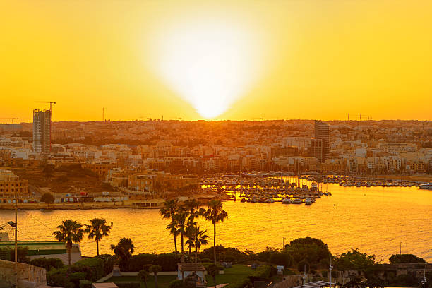 Golden light spills across Sliema harbor as boats rest beneath a fiery sunset in Malta, with palm trees framing waterfront. Warm hues bathe the skyline, blending nature and city in quiet harmony