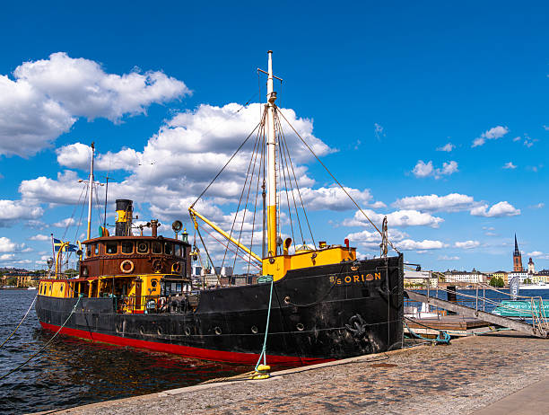 Stockholm, Sweden - 5 June 2025: A vintage tugboat named Orion docked at a harbor under a bright blue sky with fluffy clouds. The boat features a black and yellow color scheme.