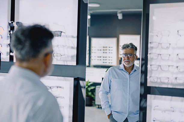 istock Senior man browsing eyeglass frames in a modern optical store. He is standing near a display wall, thoughtfully considering his options. The store has a clean, bright interior with a wide variety of eyewear on display. 2228208785