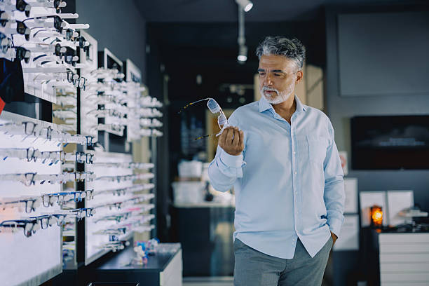 istock Senior man browsing eyeglass frames in a modern optical store. He is standing near a display wall, thoughtfully considering his options. The store has a clean, bright interior with a wide variety of eyewear on display. 2228208926