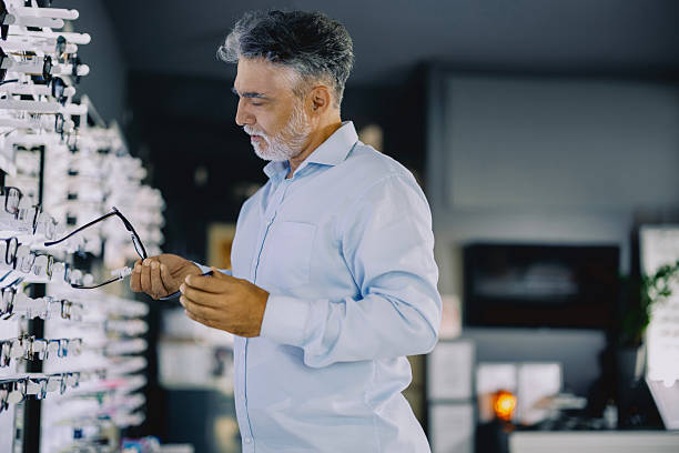 istock Senior man browsing eyeglass frames in a modern optical store. He is standing near a display wall, thoughtfully considering his options. The store has a clean, bright interior with a wide variety of eyewear on display. 2228209105