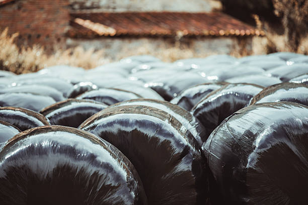Close-up view of shiny black plastic-wrapped hay bales stacked in a field near a rustic farm building with terracotta roof tiles, creating an abstract rural texture with strong light reflections