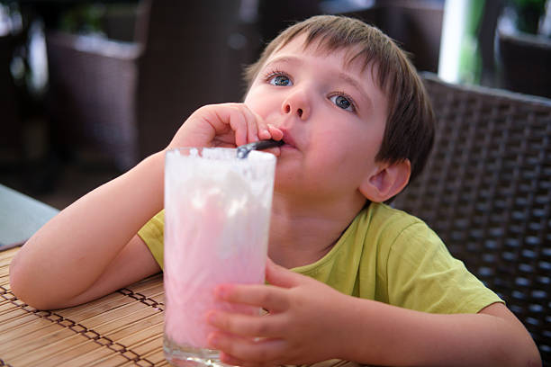 A young caucasian boy enjoying a delicious pink milkshake in a glass with a straw, sitting at a cafe table on a summer day. A child aged four years.