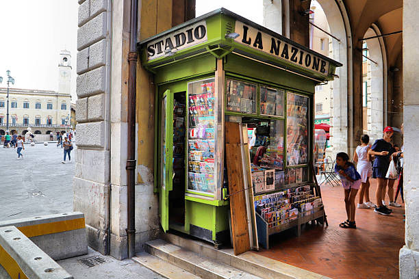 Lucca, Tuscany, Italy – September 6, 2019:  A small, vibrant green newsstand occupies a corner under arched colonnades. Its exterior displays numerous magazines and newspapers, with signs reading \