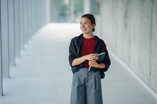 istock Smiling young woman holding notebook walking in modern office hallway 2228240628
