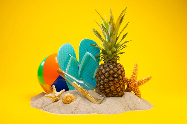 Studio shot of a colorful beach ball, flip flops, message in a bottle , starfish, and tropical pineapple sitting on a pile of sand against a bright yellow background, representing summer fun and vacation vibes.