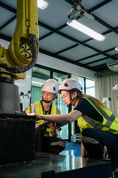 Two people wearing safety gear are looking at a machine. One of them is holding a tablet. Engineers collaborating on programming and testing automation machinery for Industry 4.0