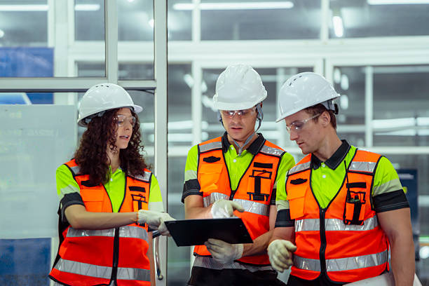 istock Three people wearing safety vests and hard hats are looking at a computer screen. Team of diverse engineers having a discussion and planning with a clipboard before operating machinery in a factory. 2228260725