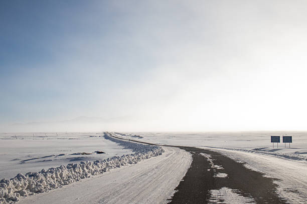 An empty winding road covered in snow and ice, surrounded by a vast snowy landscape with the horizon meeting a bright sky