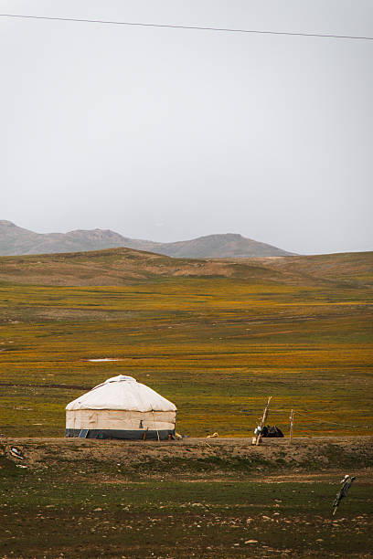 A traditional white yurt stands alone in a vast, golden and green mountain valley with hills in the background under a hazy sky