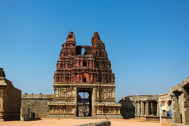 Ornate stone gateway and ruins at a historical temple complex with a vibrant red and weathered upper facade. Surrounded by stone pillars and structures, it sits on a sandy ground under a clear blue sky. Two distinct, intricately carved towers rise above the entrance. The temple is located in a historical area, reflecting ancient architectural styles and craftsmanship typical of southern India. Scattered visitors indicate the site s current use as a cultural and historical attraction.